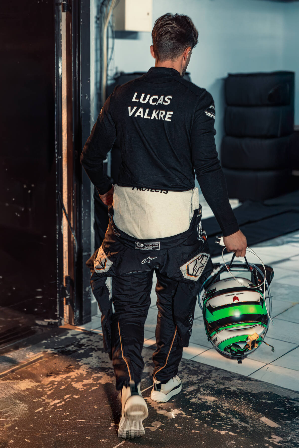 Lucas Valkre de dos, nom inscrit sur la veste, casque vert à la main, entrée du garage Krafft Racing, UCES 2025